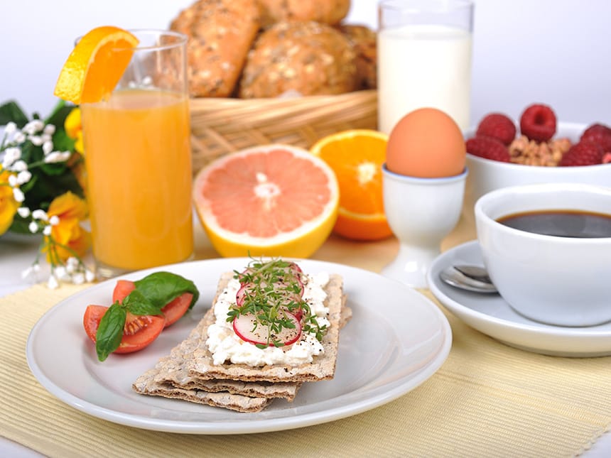 gedeckter Frühstückstisch mit Kaffe, O-Saft, Ei, Brötchen und Obst